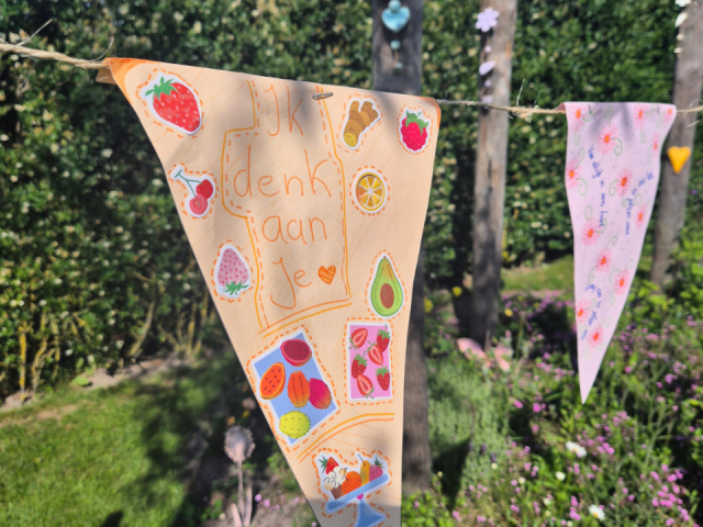 Versierd oranje vlaggetje met de tekst "Ik denk aan je", fruitstickers en een hartje, opgehangen in de tuin.